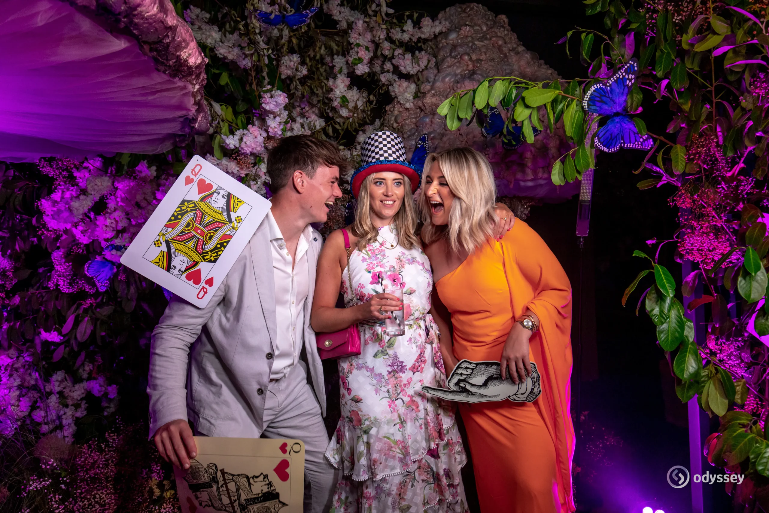Three guests enjoying an Alice in Wonderland-themed photobooth experience with oversized playing cards and colourful floral scenery.