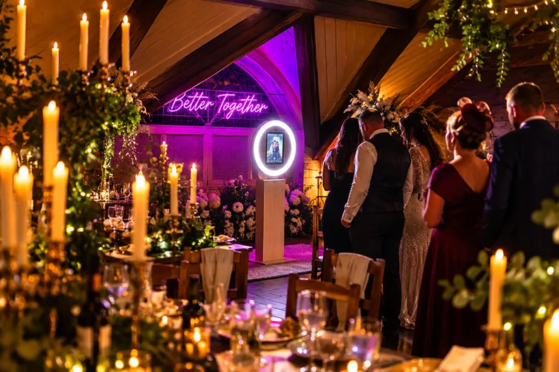 Wedding guests gathered around a photobooth in a beautifully decorated venue with warm candlelight, rustic wooden beams and a glowing Better Together neon sign